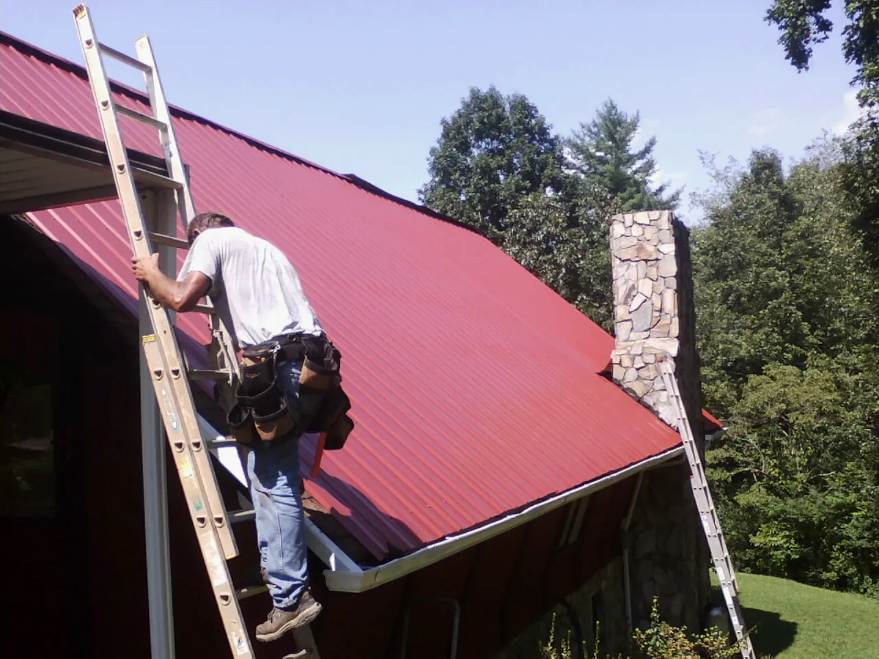 Roofer on a ladder beside a red metal roof and stone chimney — Soffit & Fascia Repair by Roofing Brothers in Canyon Creek